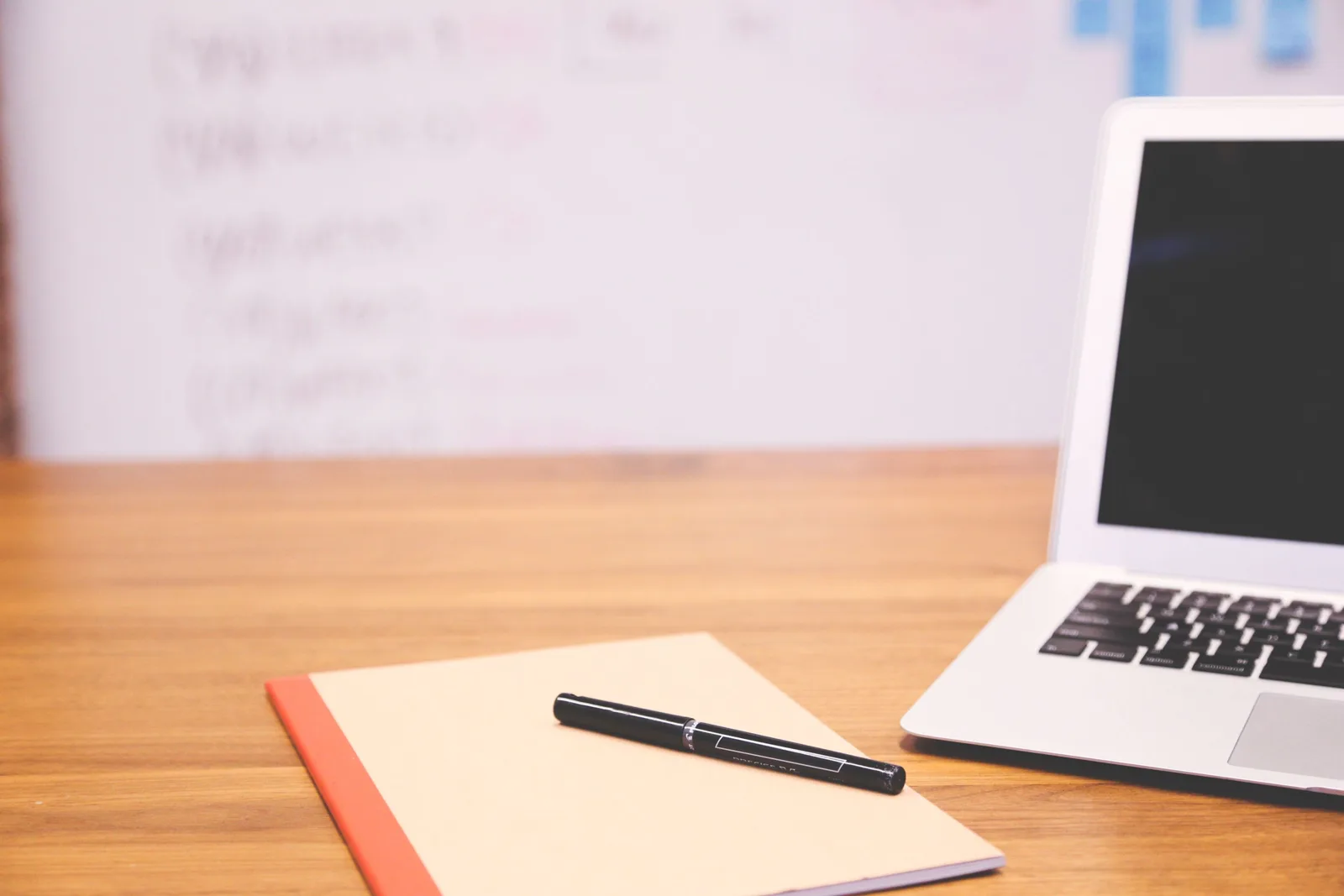 Laptop, pen and notebook on a wooden desk with a whiteboard behind